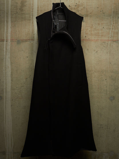 A black sleeveless long coat with a high collar and asymmetrical front opening, hanging against a textured concrete wall.