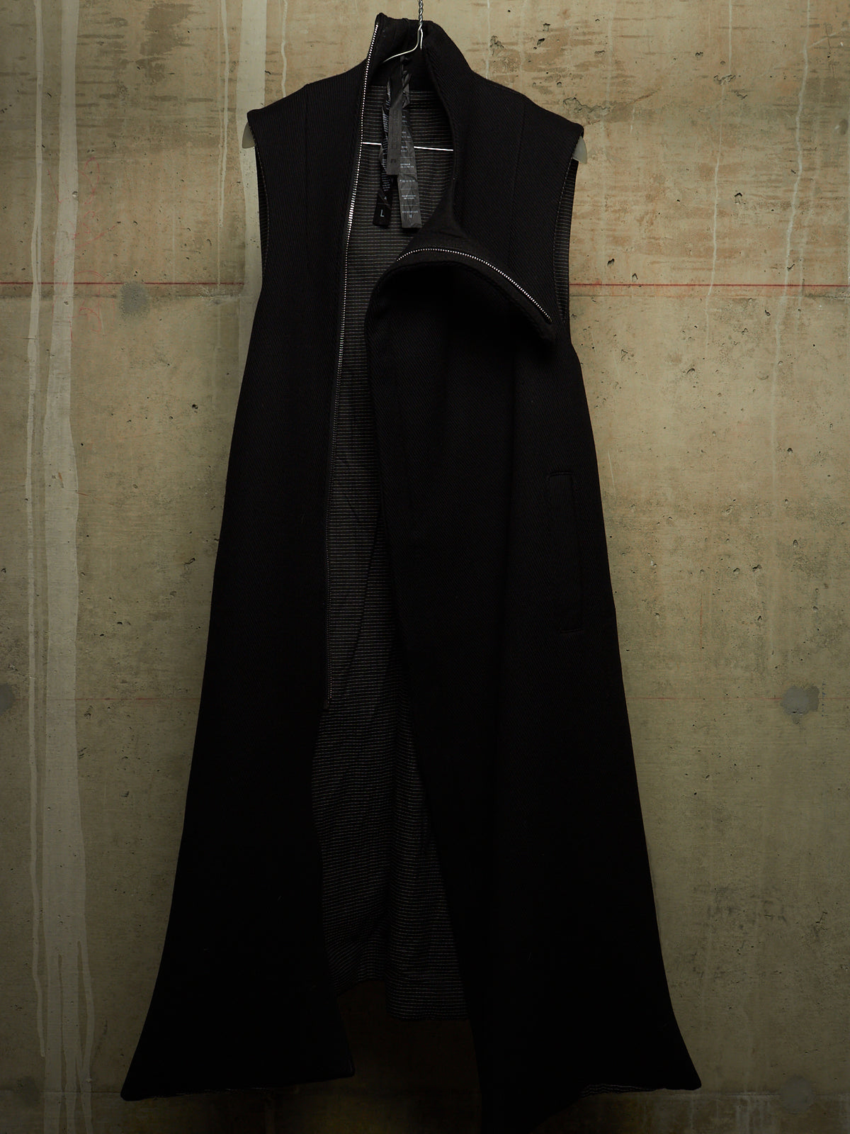 A long black sleeveless coat with a high collar and front zipper, hanging against a textured concrete wall.