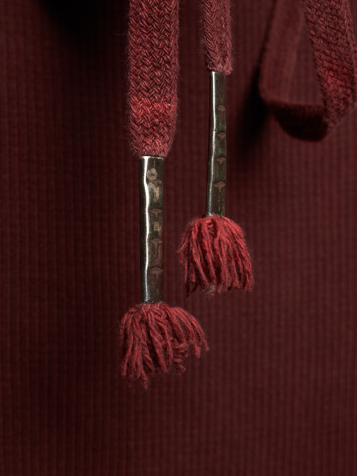 Close-up of a dark burgundy ribbed knit garment featuring textured drawstrings with metallic silver-toned shafts and matching burgundy tassels.