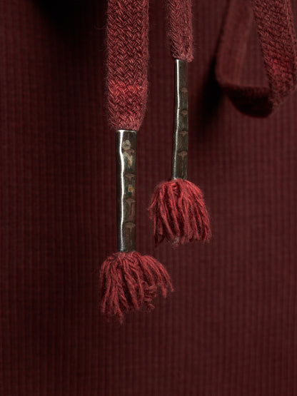 Close-up of a dark burgundy ribbed knit garment featuring textured drawstrings with metallic silver-toned shafts and matching burgundy tassels.