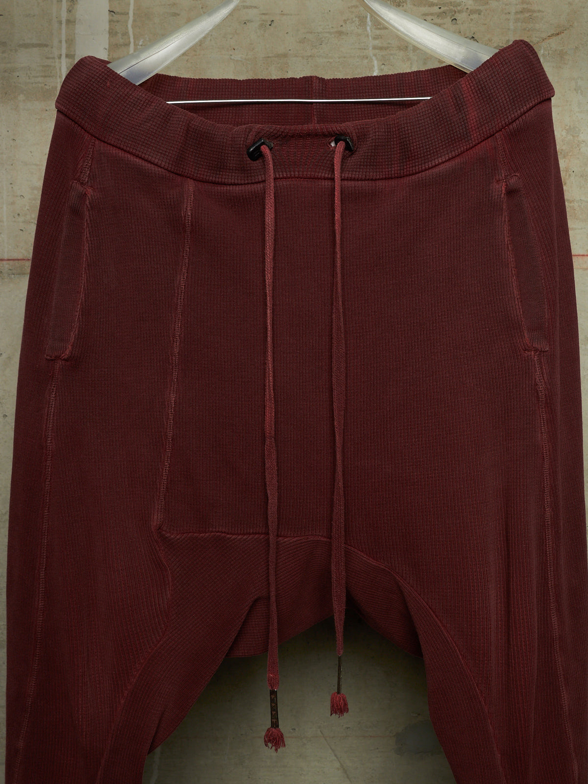 A pair of dark burgundy corduroy pants with a drawstring waist, hanging on a white hanger against a textured beige wall.