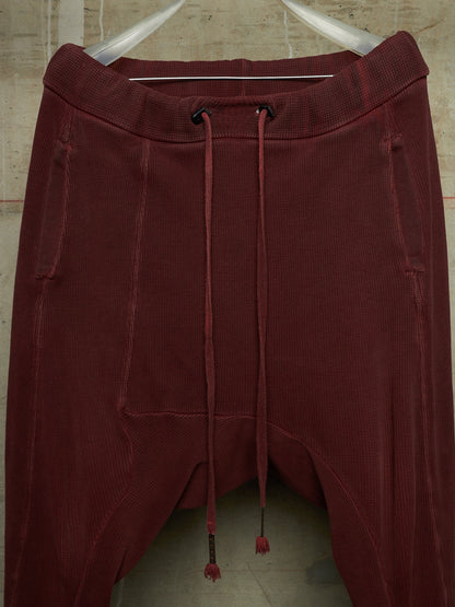 A pair of dark burgundy corduroy pants with a drawstring waist, hanging on a white hanger against a textured beige wall.