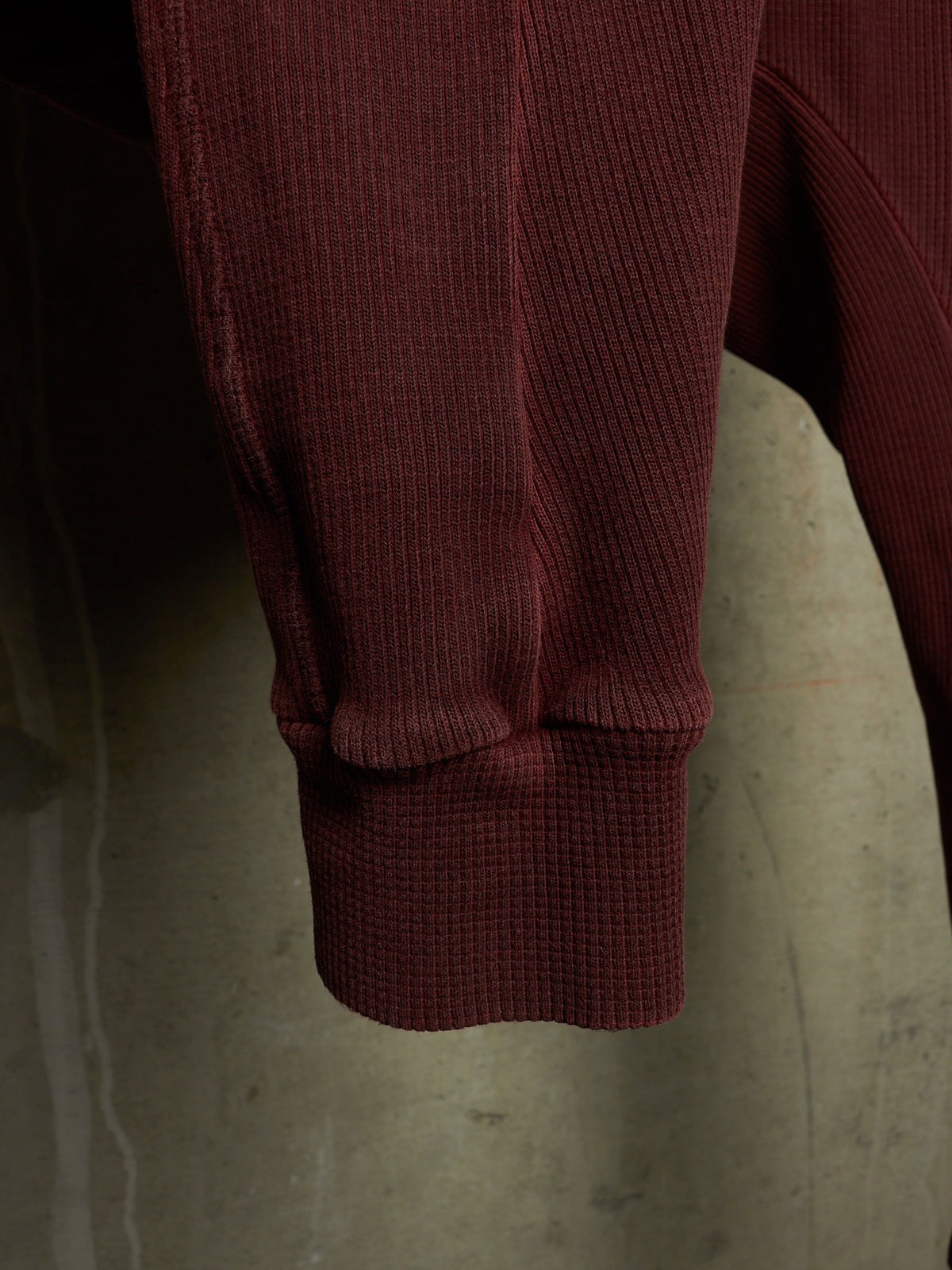 Close-up of a ribbed knit garment sleeve in a deep burgundy or maroon color, hanging against a neutral concrete-textured background.