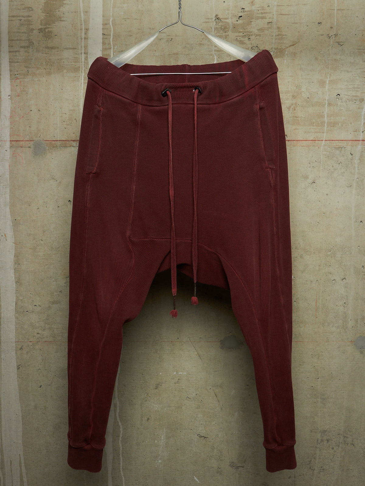A pair of dark red harem-style sweatpants hanging from a white plastic hanger against a textured concrete wall.