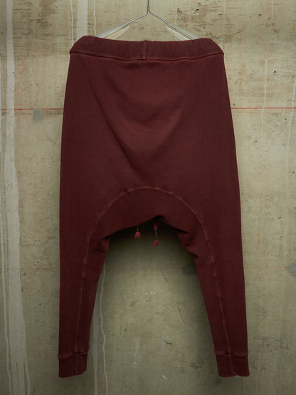 A pair of dark burgundy harem-style pants hanging from a metal hanger against a textured concrete wall.