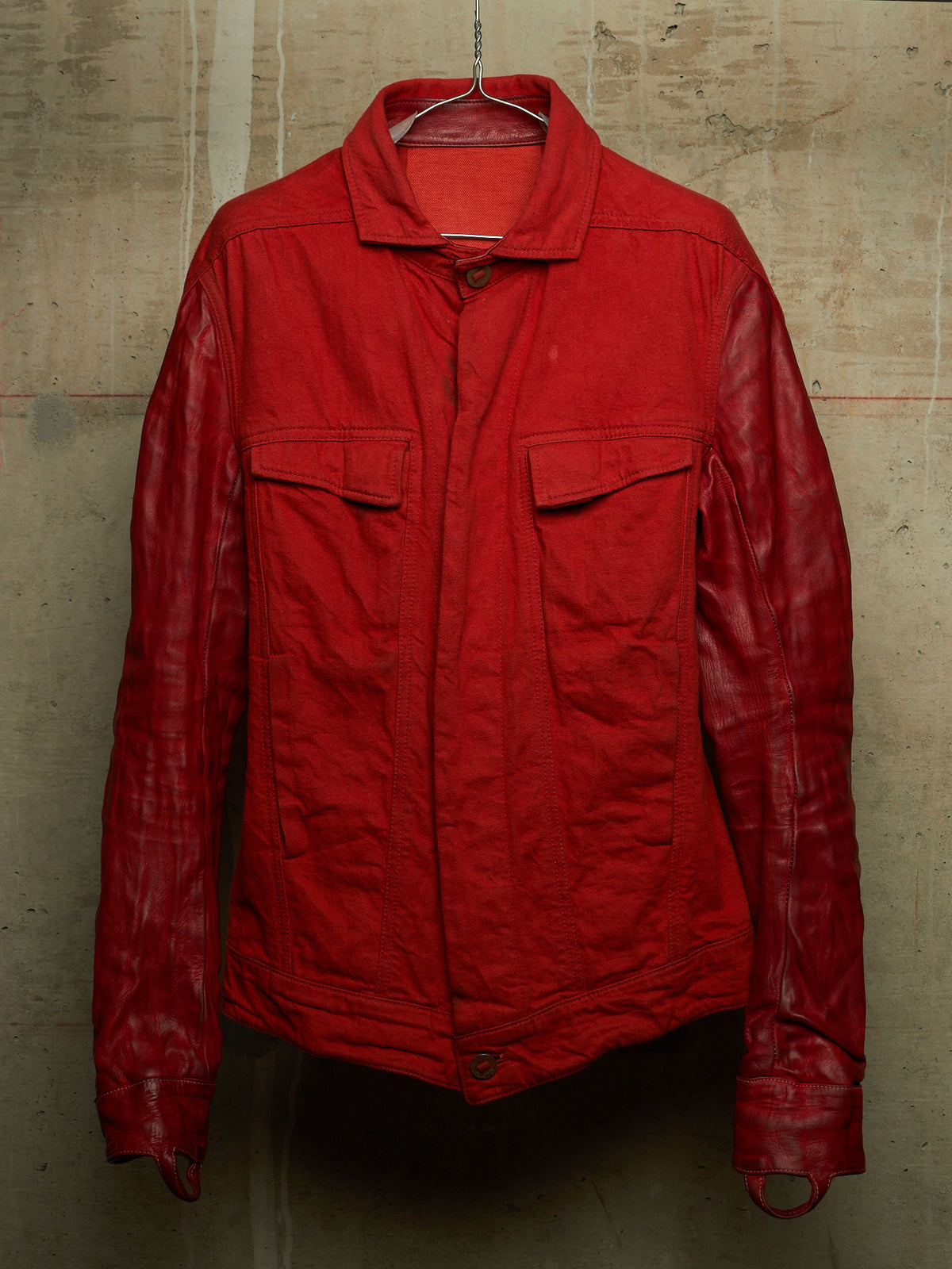 A red jacket with a matte textured body and glossy leather sleeves, hanging on a wire hanger against a concrete wall.
