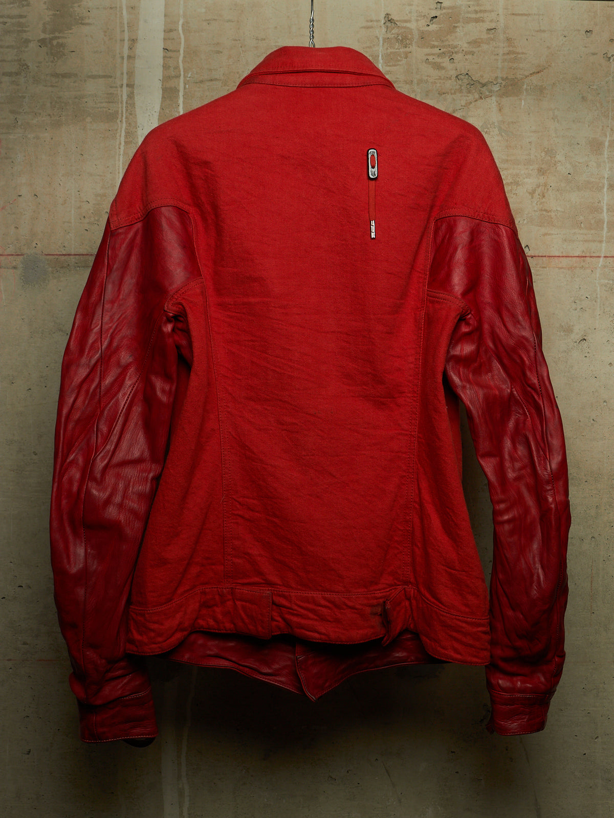 A red jacket with a matte red body and glossy red leather sleeves, shown from the back against a textured concrete wall.