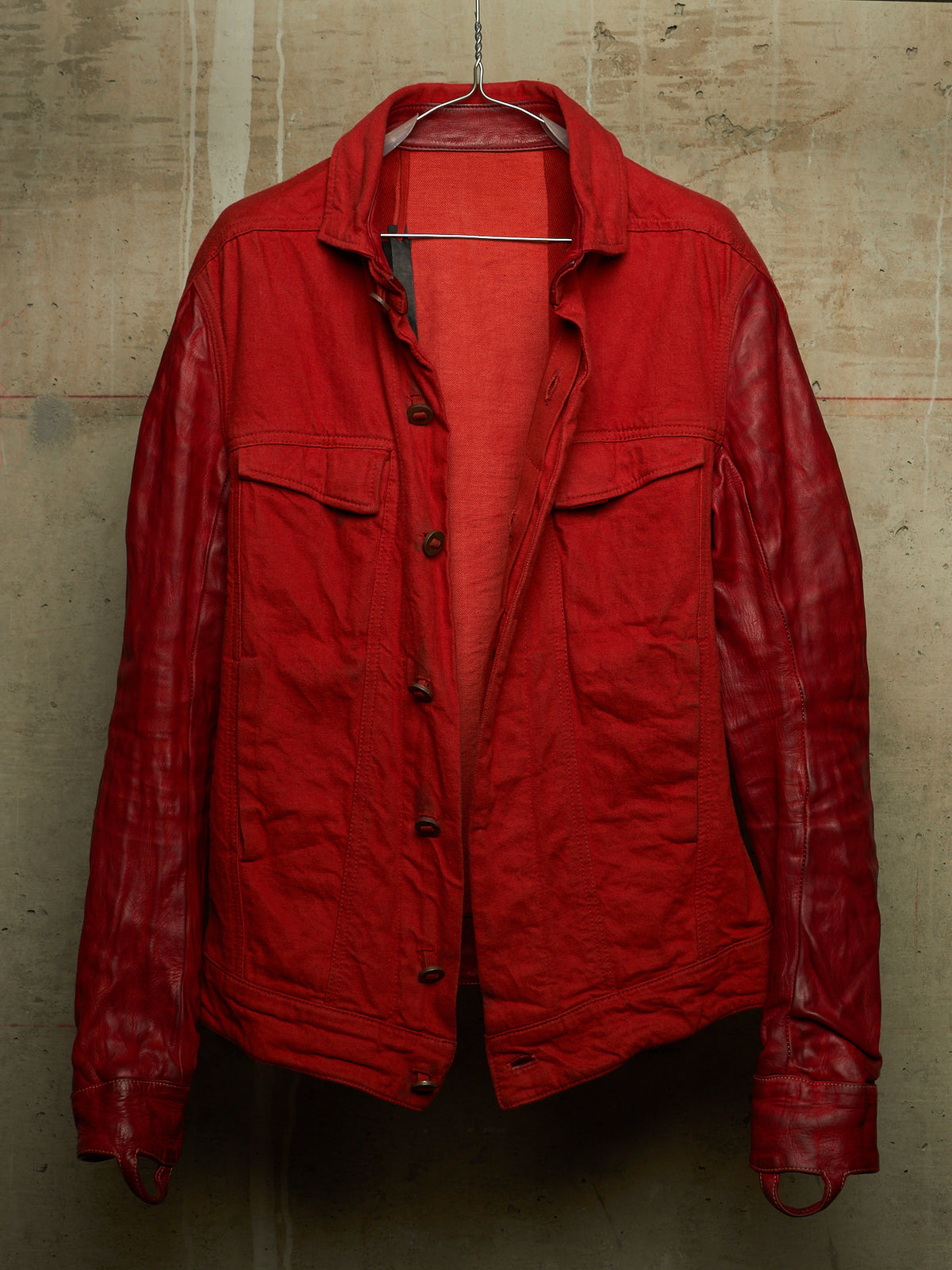 A red jacket with a matte cotton body and glossy leather sleeves, hanging on a wire hanger against a textured concrete wall.