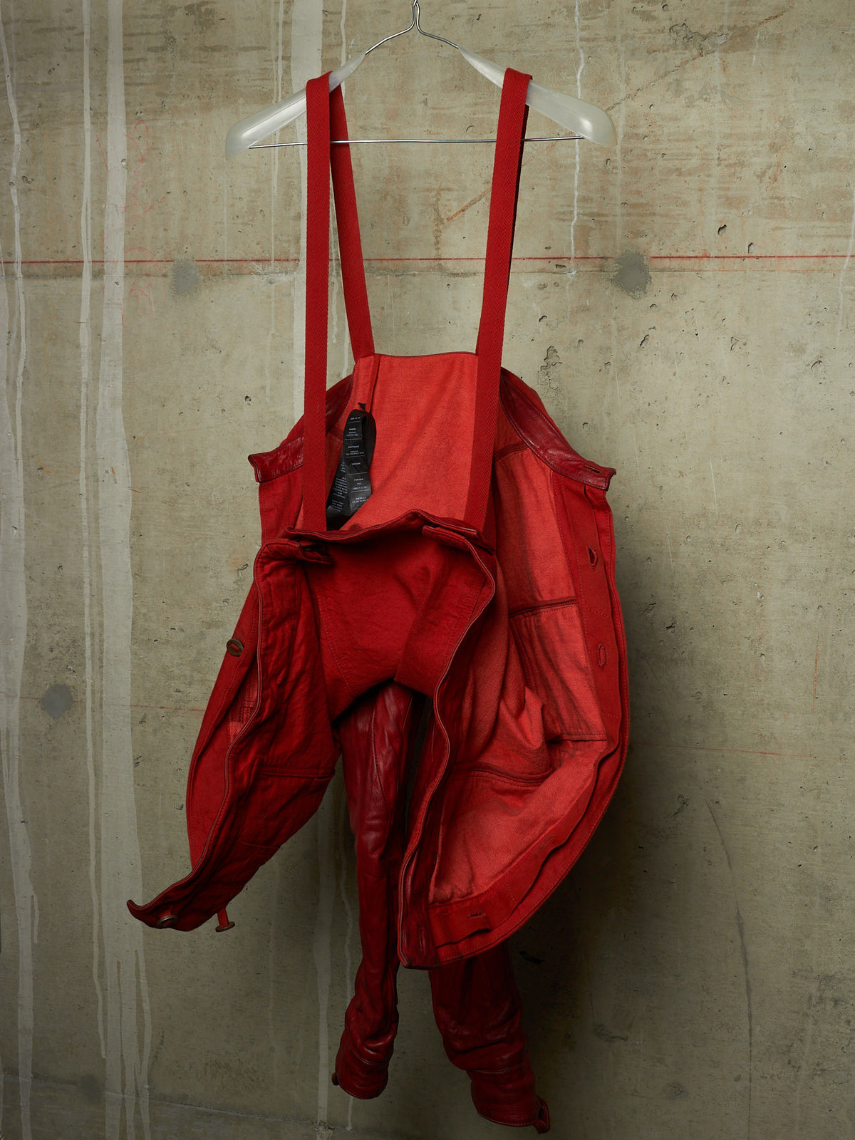 A bright red waterproof waders suit with shoulder straps, hanging from a clear plastic hanger against a textured concrete wall.
