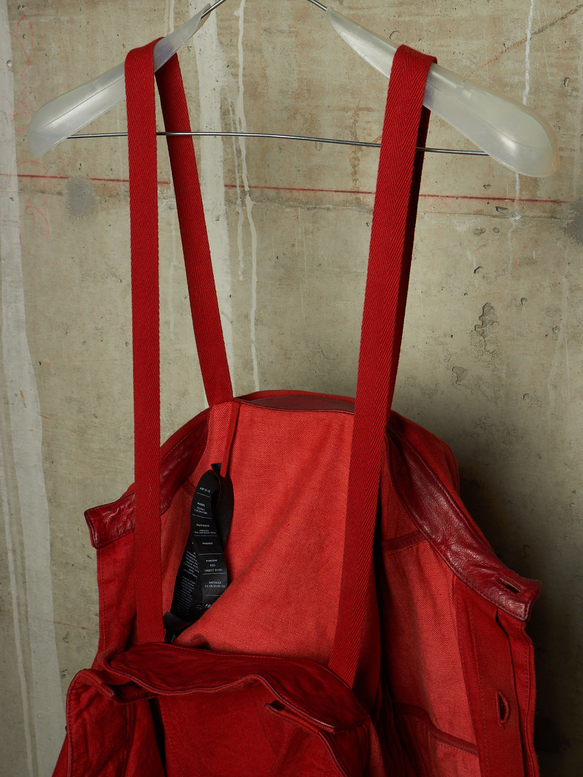 A red canvas tote bag with wide shoulder straps hanging from a clear plastic hanger against a textured concrete wall.