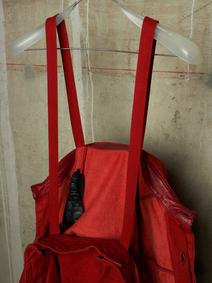 A red canvas tote bag with wide shoulder straps hanging from a clear plastic hanger against a textured concrete wall.