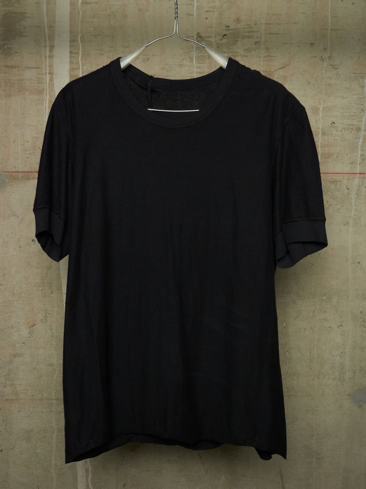 A black short-sleeve crewneck t-shirt hanging on a metal hanger against a textured concrete wall.