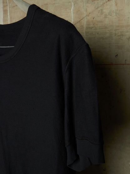 A close-up of a black short-sleeve knit top hanging against a textured concrete wall.