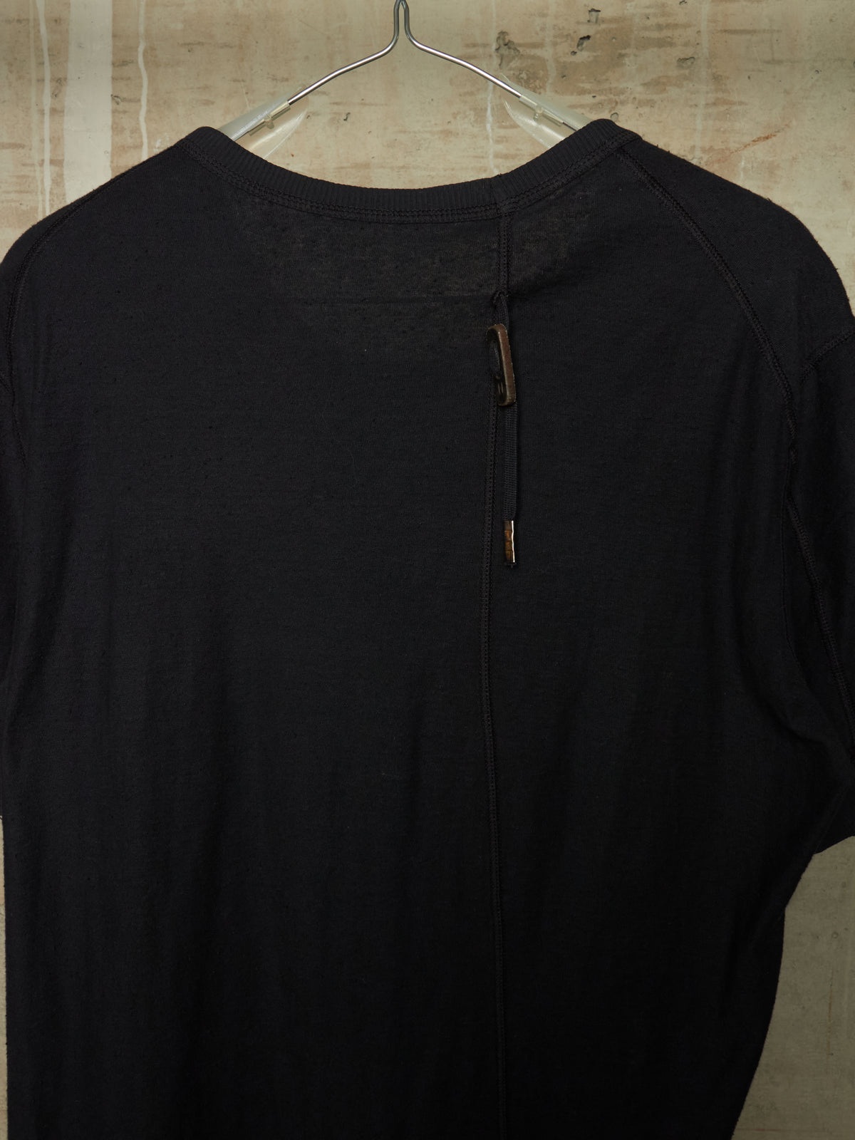 A black knit long-sleeve top hanging on a clear plastic hanger against a textured beige wall. The back of the shirt features a central vertical seam with two small brown leather-like pull tabs.