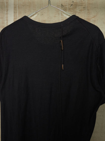 A black knit long-sleeve top hanging on a clear plastic hanger against a textured beige wall. The back of the shirt features a central vertical seam with two small brown leather-like pull tabs.