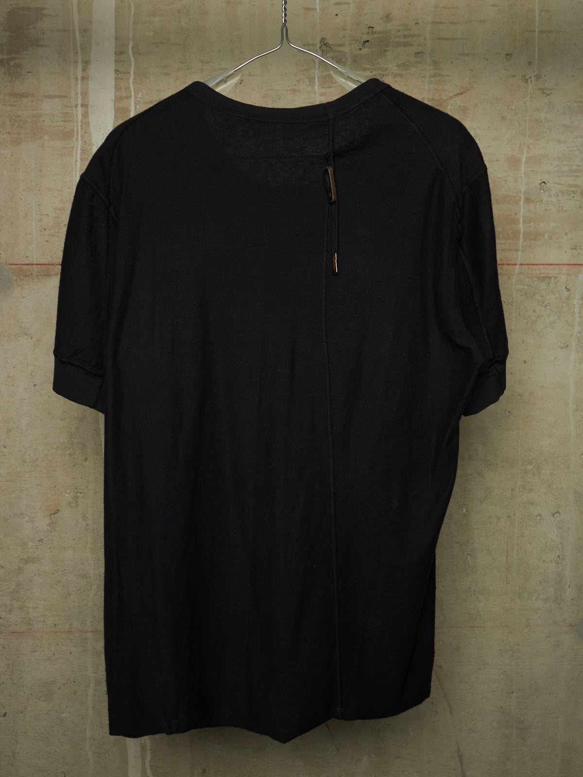 A black short-sleeve top hanging on a metal hanger against a textured concrete wall. The shirt features a unique front zipper closure with small brown pull tabs and a sheer mesh yoke at the neckline.