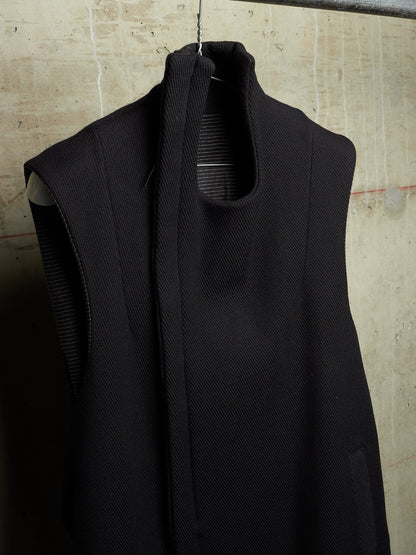 A sleeveless black textured knit vest with a high mock neck and a wide front tie detail, hanging against a textured concrete wall.