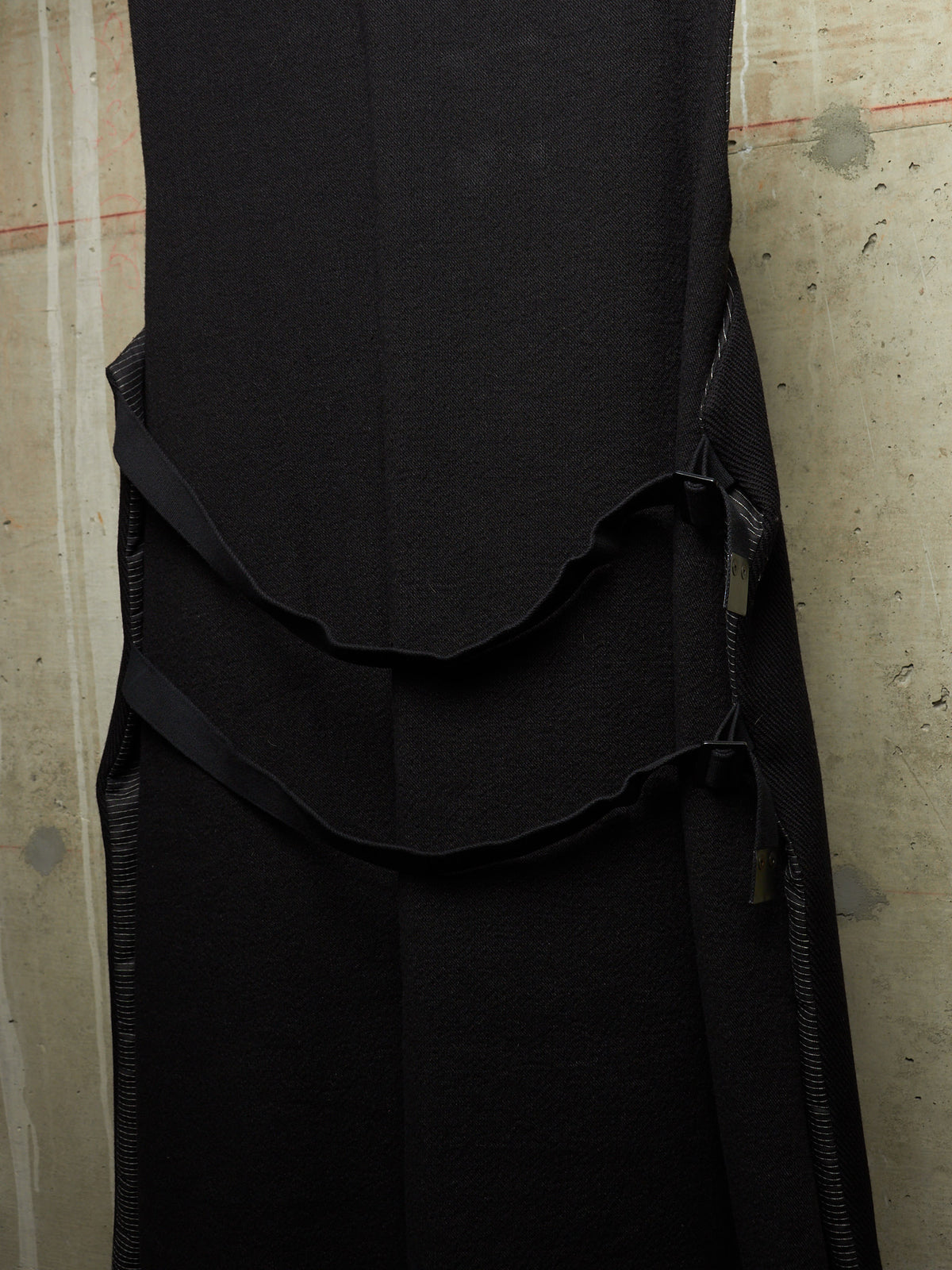 A close-up of a black sleeveless dress or long top with a structured bodice, layered ruffle detail, and side zipper, shown against a textured concrete wall.
