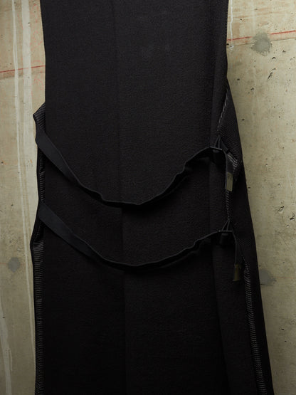 A close-up of a black sleeveless dress or long top with a structured bodice, layered ruffle detail, and side zipper, shown against a textured concrete wall.
