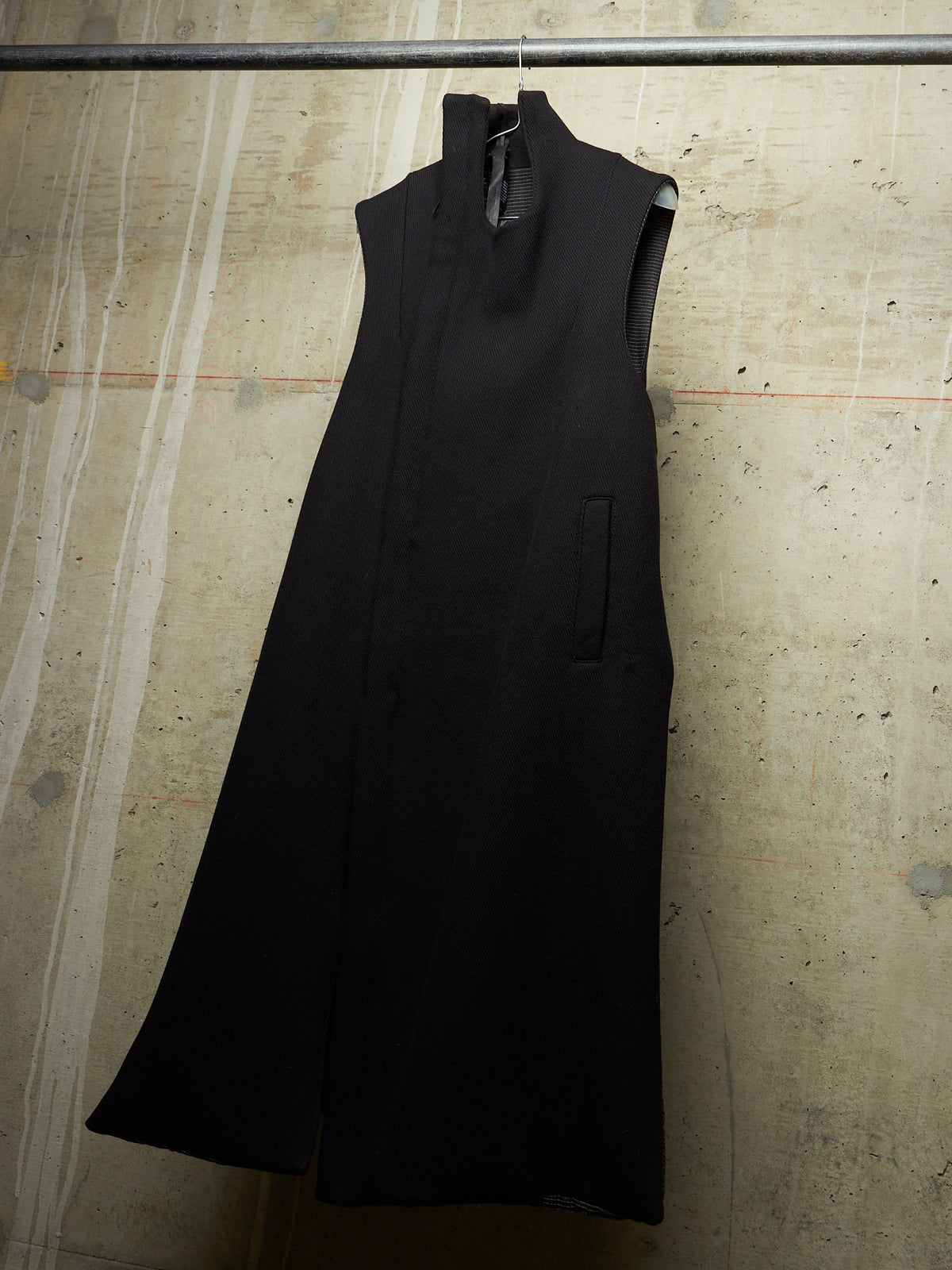 A sleeveless black high-neck dress or long coat hanging on a hanger against a textured concrete wall.