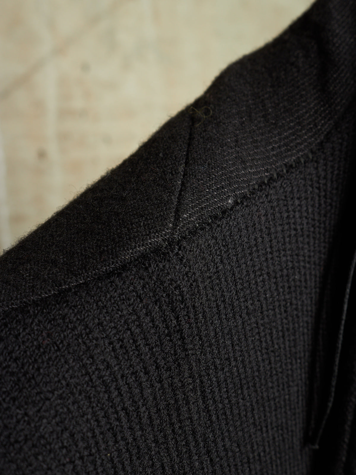 Close-up detail of a black ribbed knit garment, showing the texture and stitching of the collar area against a light beige background.