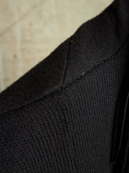 Close-up detail of a black ribbed knit garment, showing the texture and stitching of the collar area against a light beige background.