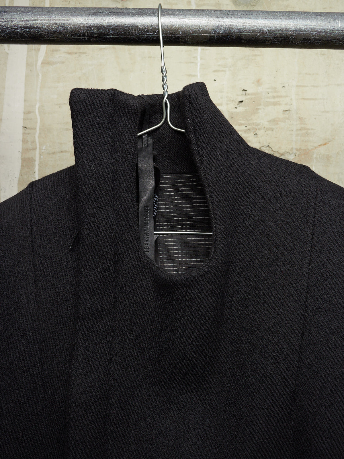 A close-up of a black textured knit garment hanging on a metal hanger from a dark horizontal rod, with a light beige stone or tile wall in the background.