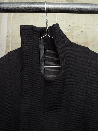 A close-up of a black textured knit garment hanging on a metal hanger from a dark horizontal rod, with a light beige stone or tile wall in the background.