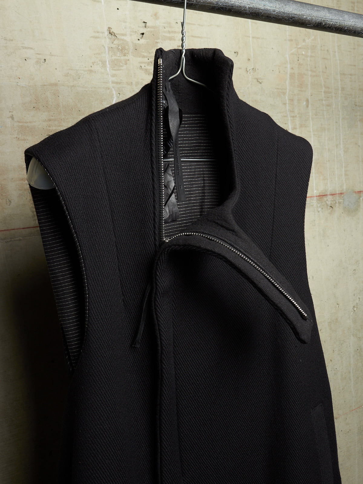 A black sleeveless high-neck jacket with a wide collar and asymmetrical zipper detail, hanging on a metal hanger against a textured concrete wall.