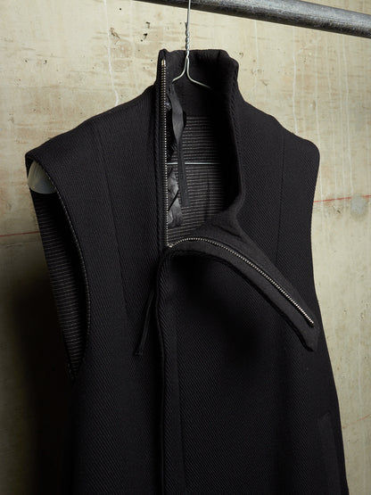 A black sleeveless high-neck jacket with a wide collar and asymmetrical zipper detail, hanging on a metal hanger against a textured concrete wall.