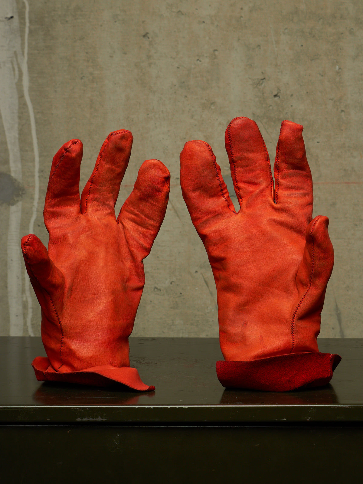 A pair of bright red leather gloves displayed upright on a dark surface against a textured concrete wall.