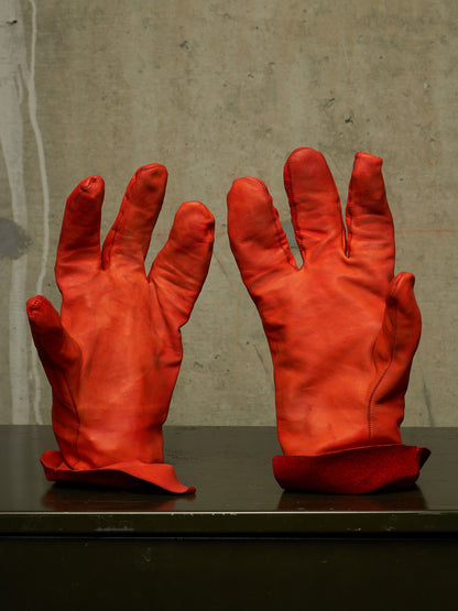 A pair of bright red leather gloves displayed upright on a dark surface against a textured concrete wall.