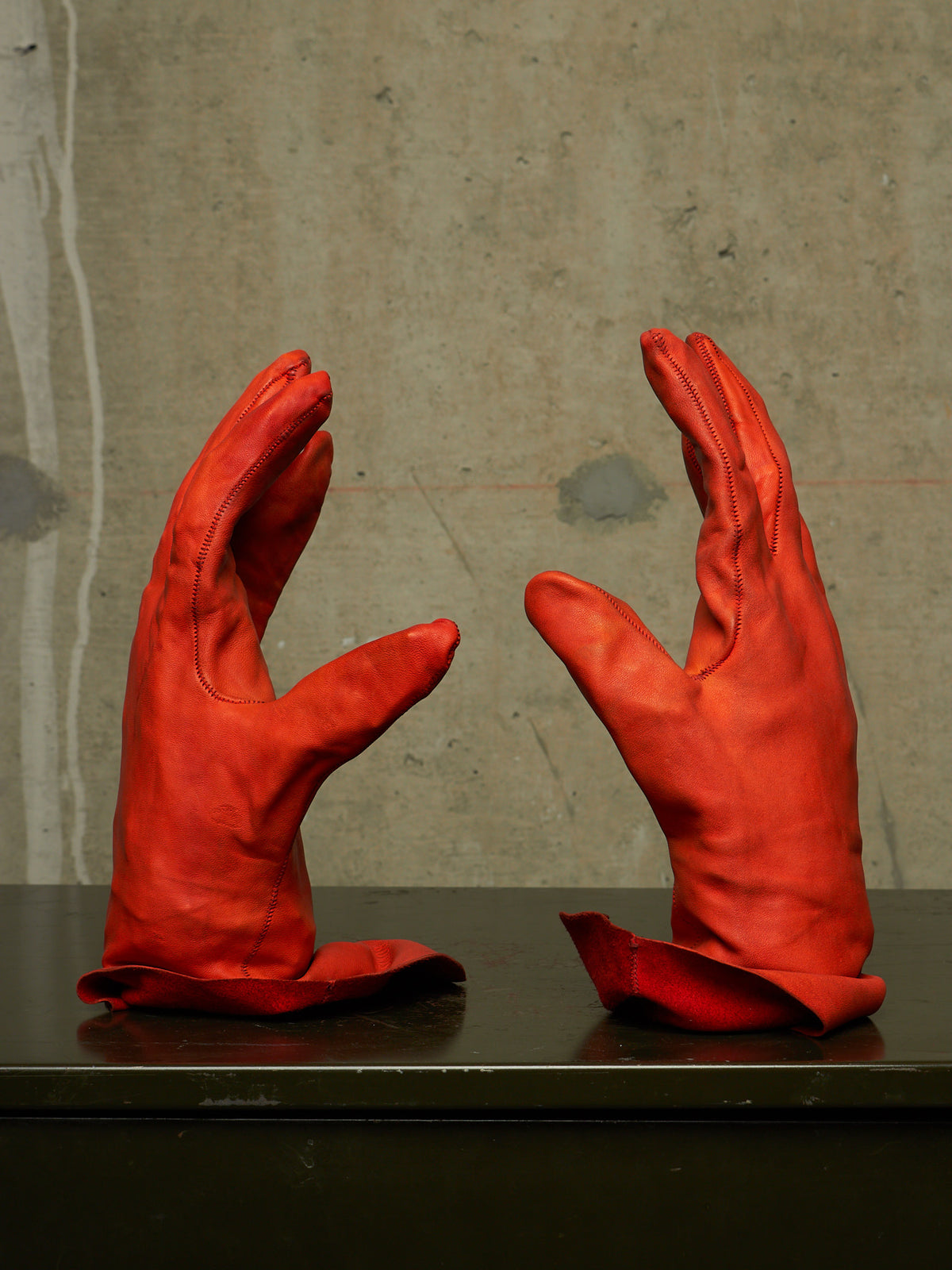 A pair of bright red leather gloves displayed upright on a dark surface against a textured concrete wall.