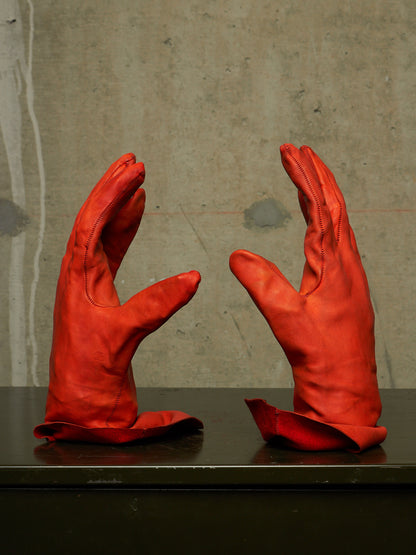 A pair of bright red leather gloves displayed upright on a dark surface against a textured concrete wall.