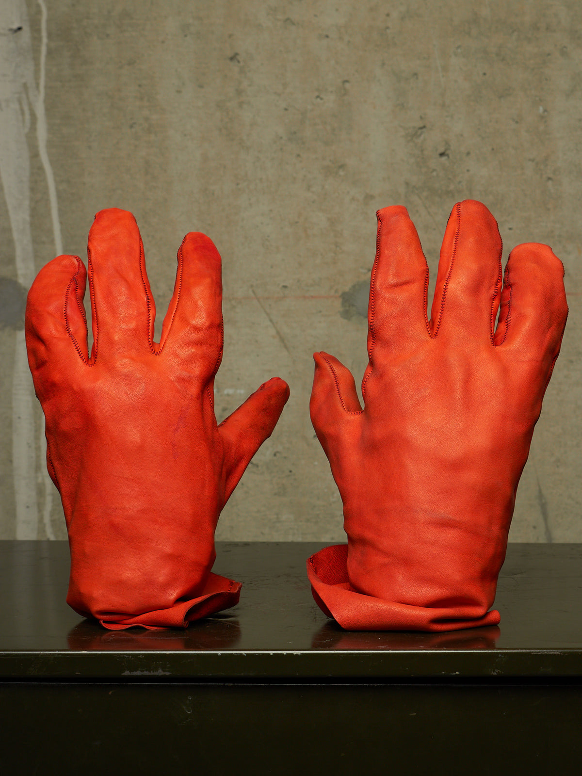 A pair of bright red leather gloves displayed side-by-side on a dark surface against a textured gray concrete wall.