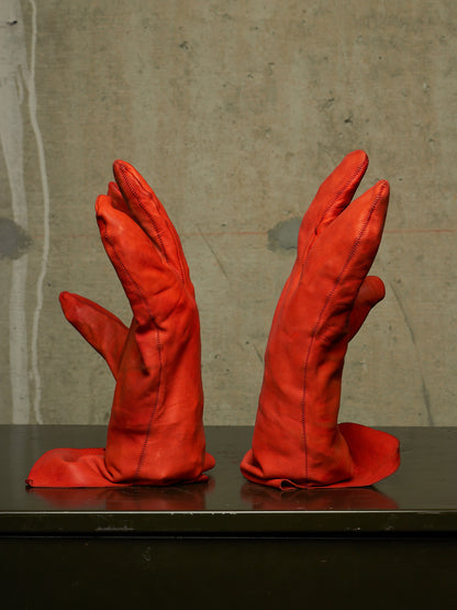 A pair of bright red leather gloves displayed upright on a dark reflective surface against a textured gray concrete wall.