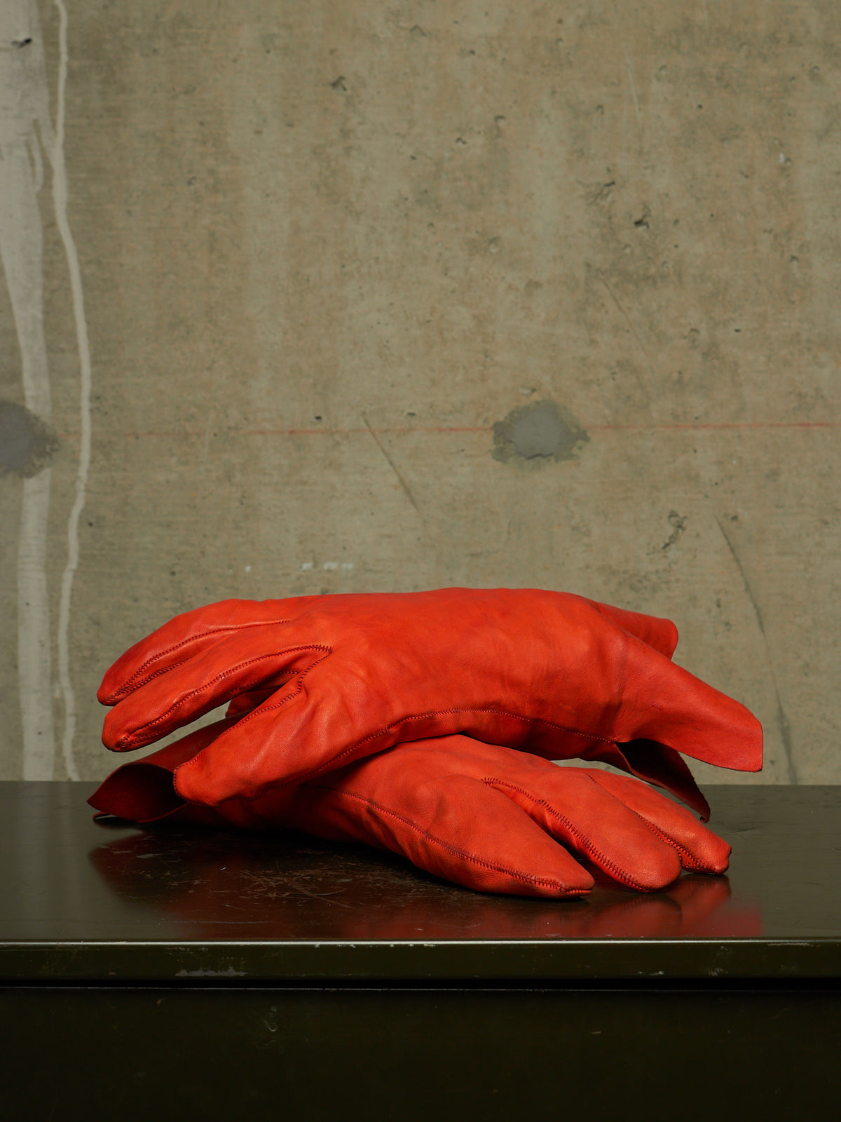 A pair of bright red leather gloves stacked on a dark reflective surface against a textured concrete wall.
