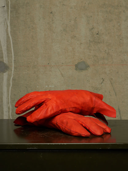 A pair of bright red leather gloves stacked on a dark reflective surface against a textured concrete wall.