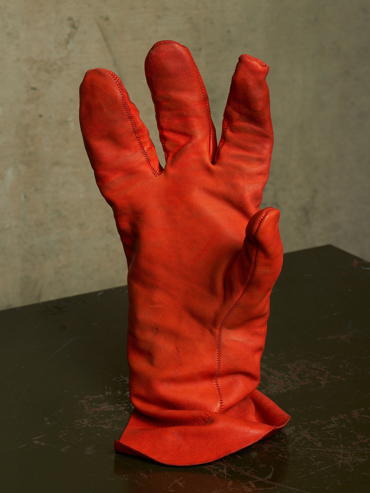 A single red leather protective glove standing upright on a dark surface against a textured gray wall.