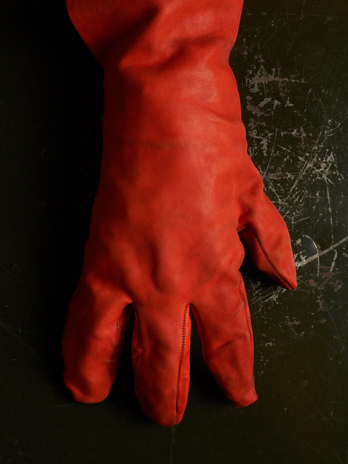 A single long red leather glove laid flat on a dark textured surface.