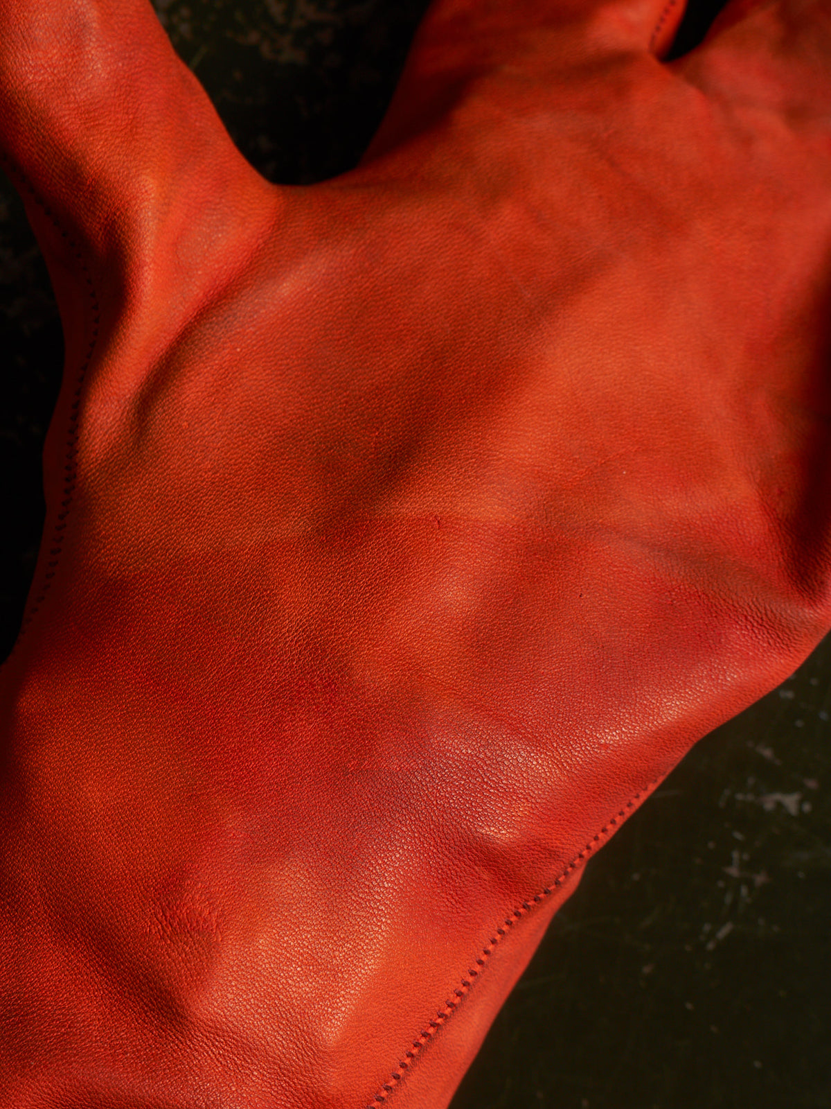 A close-up of a vibrant orange-red leather glove, showing the smooth texture and fine stitching against a dark background.