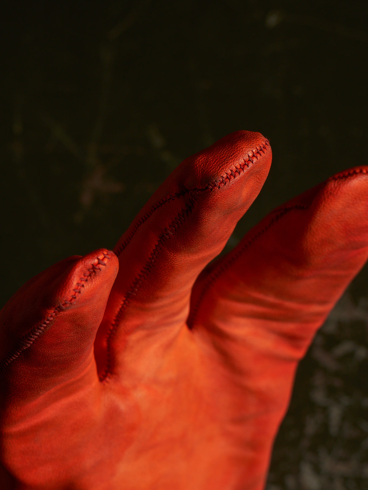 A close-up of a red leather glove or mitten with visible finger seams, photographed against a very dark background.