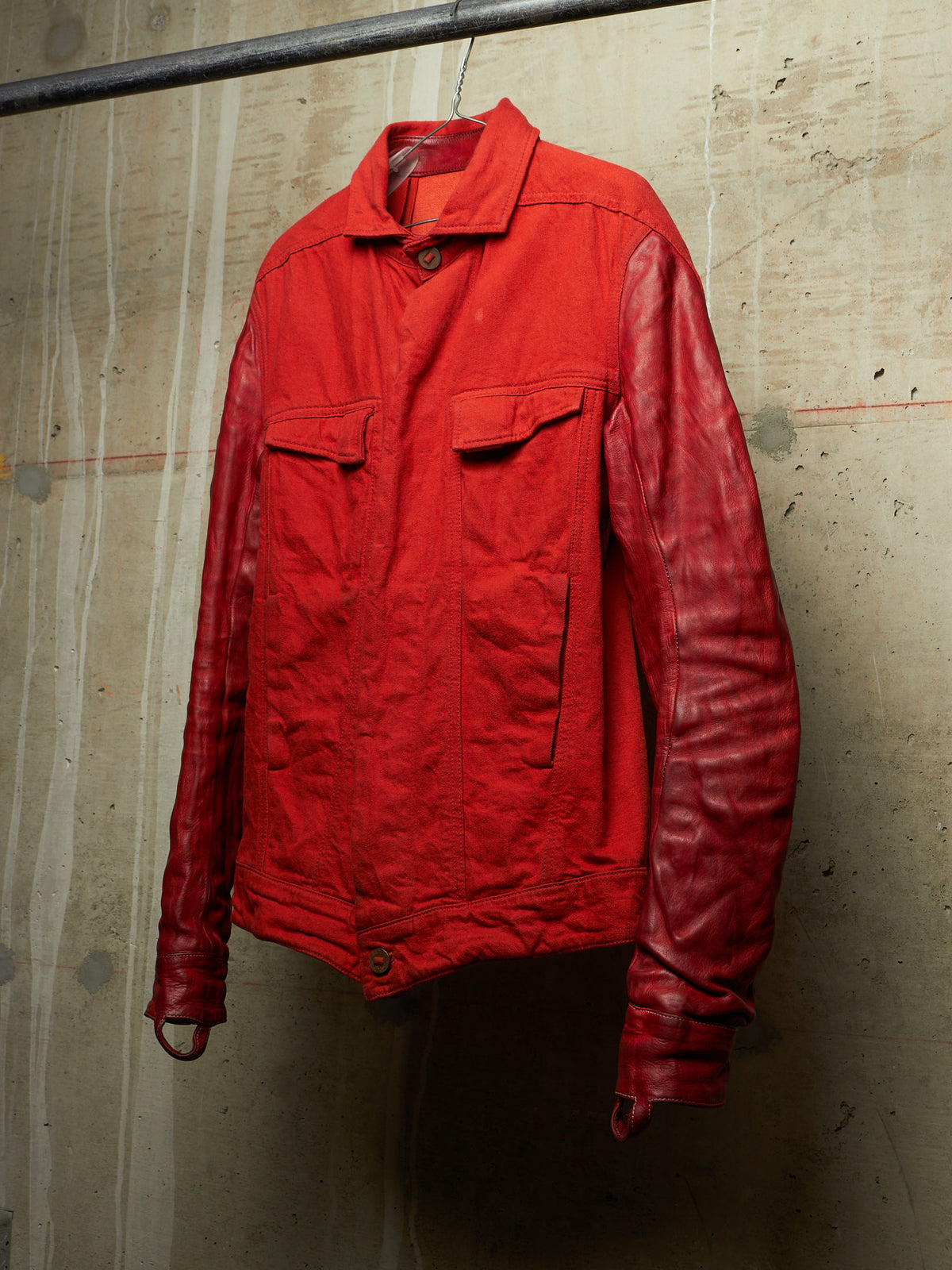 A red jacket with a matte red body and glossy red sleeves, hanging on a metal hanger against a textured concrete wall.