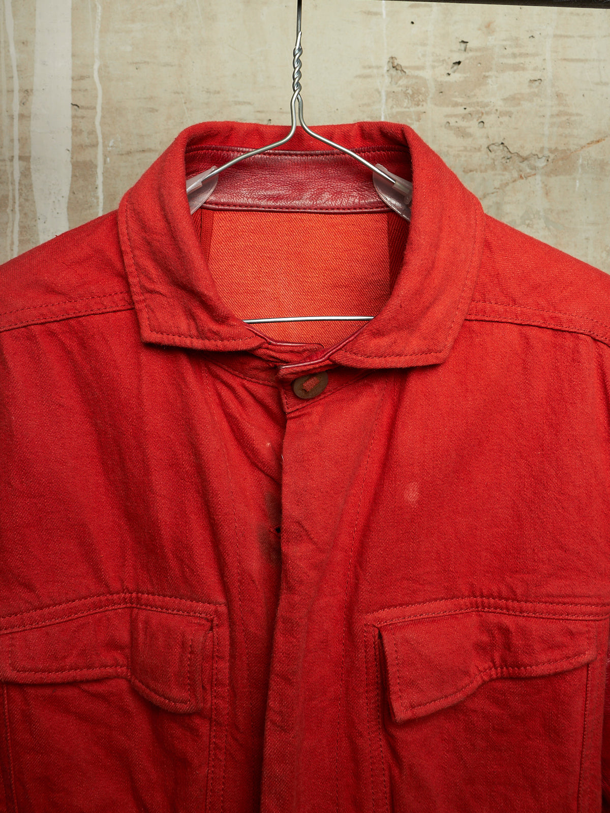 A red canvas utility jacket with a pointed collar and button closure, hanging on a wire hanger against a textured beige wall.