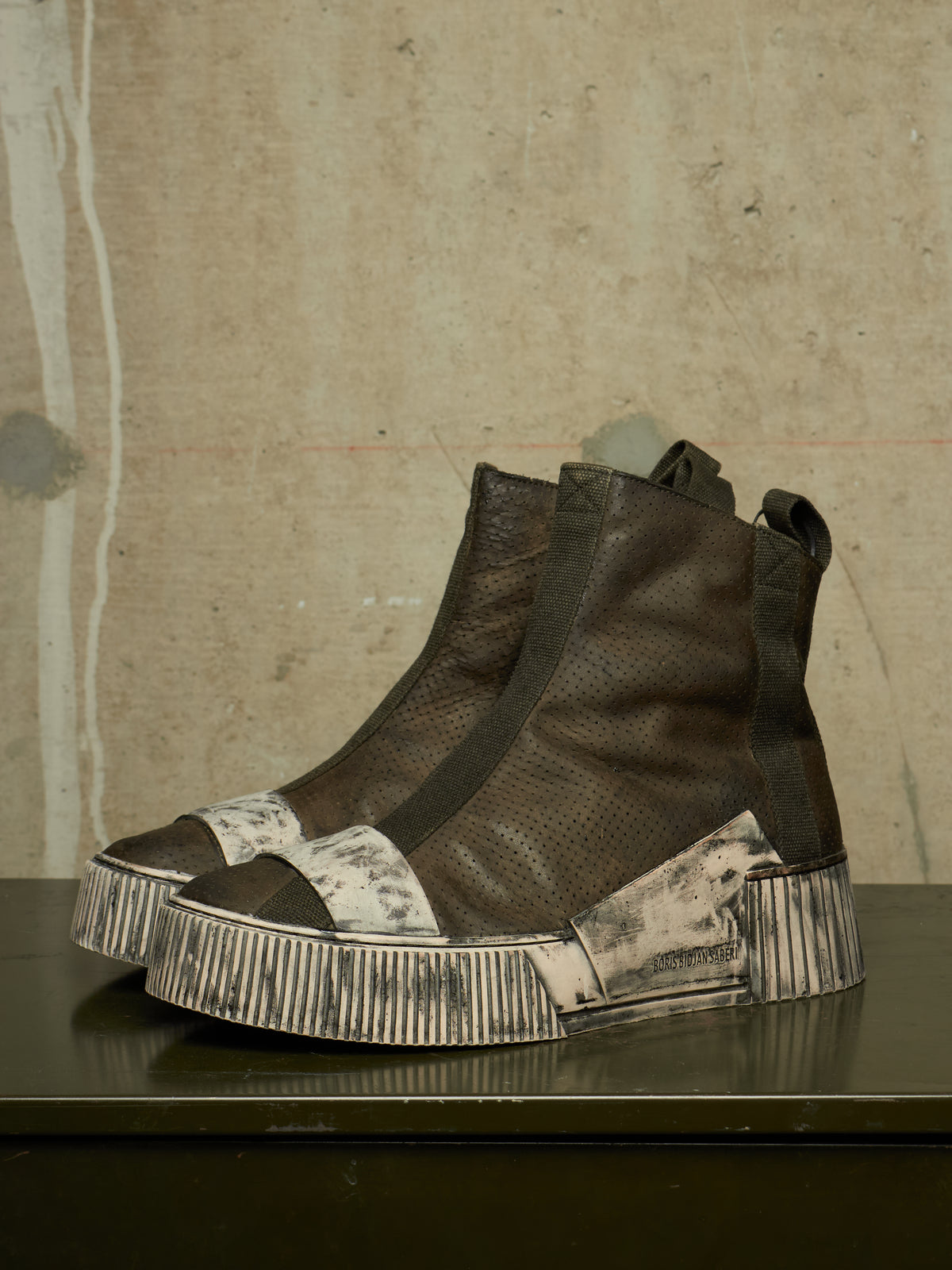A pair of high-top ankle boots with dark brown textured leather uppers, wide black elastic side panels, and distressed off-white platform soles, displayed on a dark reflective surface against a textured concrete wall.
