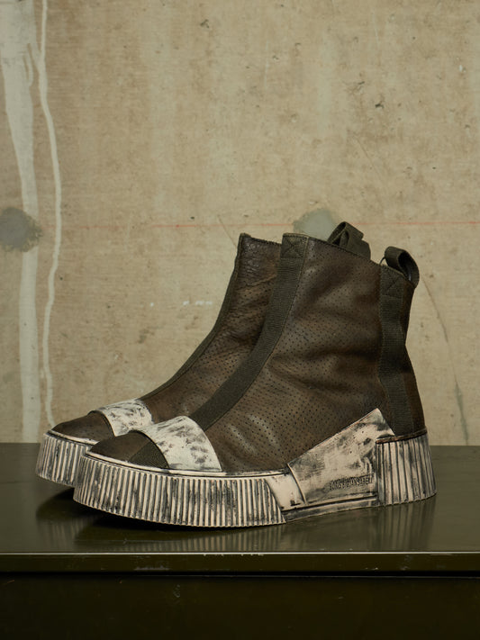 A pair of high-top ankle boots with dark brown textured leather uppers, wide black elastic side panels, and distressed off-white platform soles, displayed on a dark reflective surface against a textured concrete wall.
