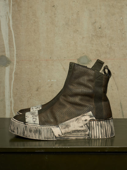 A single high-top sneaker boot with distressed black leather and a heavily weathered, off-white sole, displayed on a dark table against a textured concrete wall.