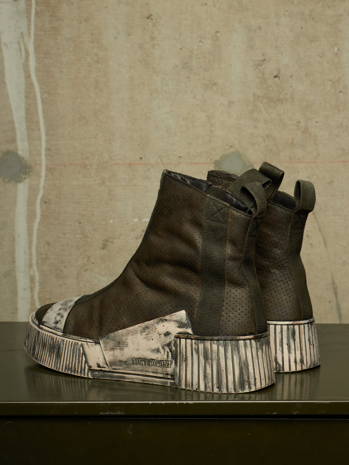 A pair of dark brown textured leather ankle boots with chunky, distressed silver and black platform soles, displayed on a dark table against a textured concrete wall.