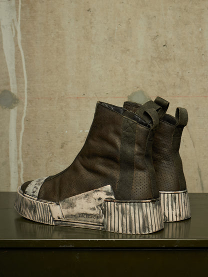 A pair of dark brown textured leather ankle boots with chunky, distressed silver and black platform soles, displayed on a dark table against a textured concrete wall.