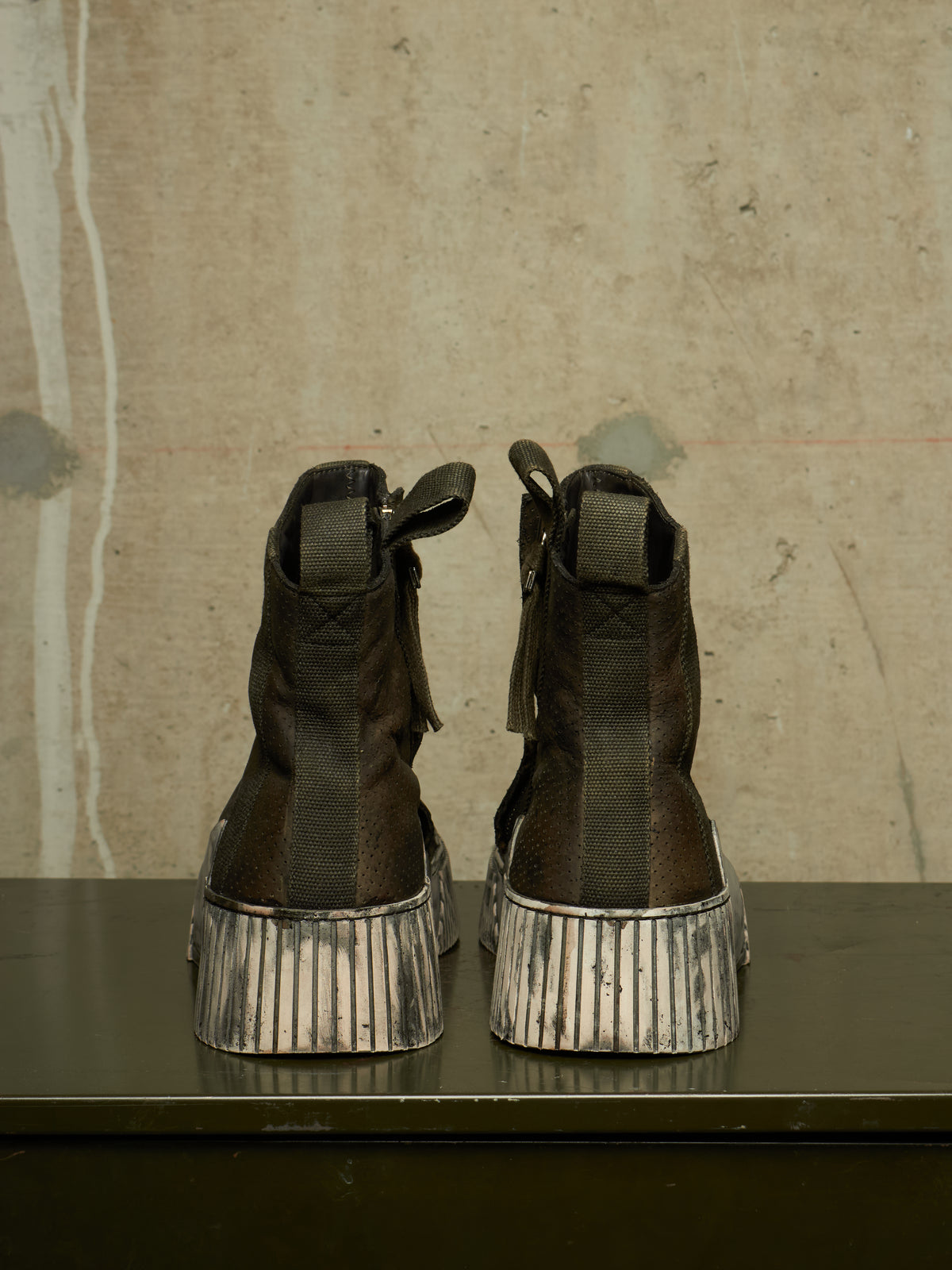 A pair of dark brown leather ankle boots with distressed silver platform soles, shown from the back on a dark table against a textured concrete wall.